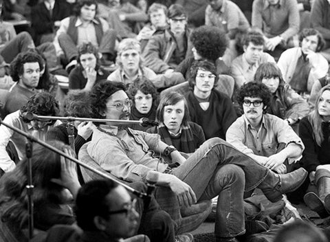 A group of people, predominantly young adults, are sitting closely together on the floor, facing toward the left side of the frame. In the foreground, a man with glasses and a mustache is seated, leaning forward slightly and speaking into a microphone. He is wearing a collared shirt and jeans. Several other individuals are visible behind him, some looking towards the speaker, others with more neutral expressions. The overall scene suggests a gathering or event where a speaker is addressing an audience, like