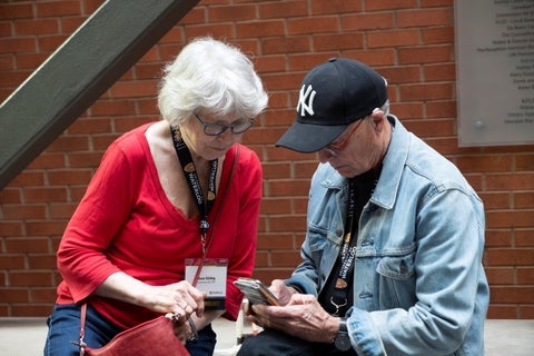 An older woman with short grey hair and glasses in a red shirt and an older man wearing a baseball cap and a denim jacket look at a smartphone together.