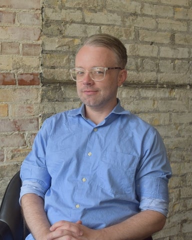 Man in glasses wearing a blue button-up shirt sits against a brick wall, posing for a portrait.