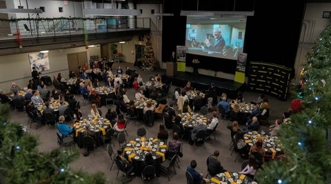 A large banquet hall decorated for the holidays with people sitting at round tables, a speaaker at a podium and a presentation playing on a large screen.