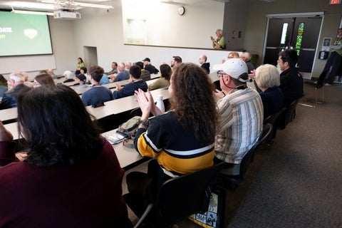 Audience in a lecture hall clapping for a speaker on a raised platform, with a projection screen displaying a logo and text in the background.