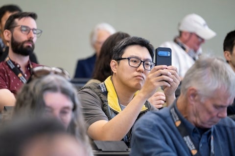 A man with glasses holds up a smartphone to take a picture or video at an indoor event, with other attendees seated in the blurred background.