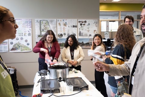 A group of individuals interacts around a table with cooking appliances and research posters in the background.
