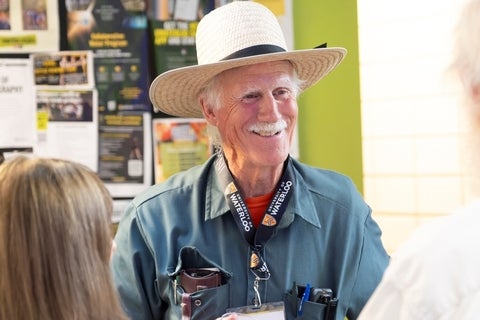 An elderly man wearing a straw hat and a teal collared shirt with a University of Waterloo lanyard smiles and speaks to someone off-camera, with a wall of posters in the background.