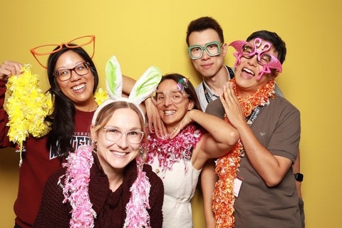 Five individuals posing for a photo booth with silly accessories against a yellow backdrop, including large novelty glasses and leis.