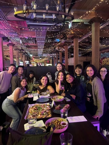 A group of friends smiles around a table laden with food and drinks in a dimly lit bar decorated with string lights and industrial-style fixtures.