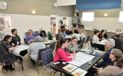 Community workshop with adults seated at tables, discussing notes and raising a hand in a bright meeting hall.