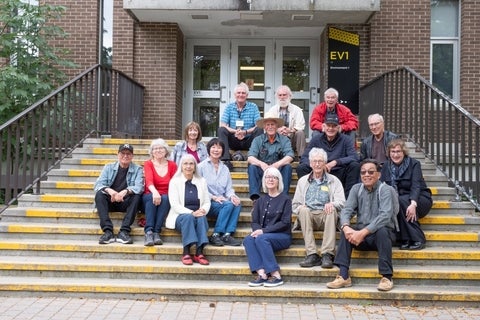 A group of people of varying ages are sitting on steps in front of a brick building with "EV1 Environment 1" signage.