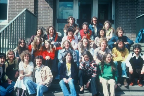 A large group of diverse individuals, appearing to be students or colleagues, pose for a candid group photo on outdoor steps in front of a brick building.