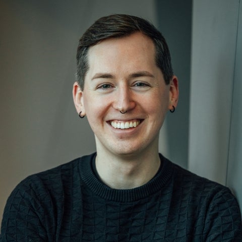 A person with short hair and a septum piercing, wearing a black sweater, smiling and leaning against a wall.