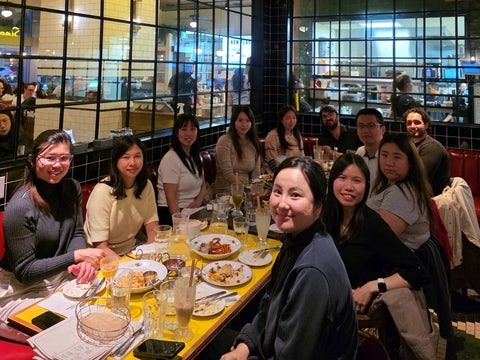 A group of people sits around a yellow table in a restaurant, with food and drinks laid out in front of them.