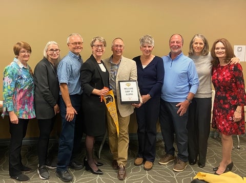 A group of nine adults stand for a photo, holding a framed certificate that reads "WELCOME SURP '81 ALUMNI" and a yellow pennant with a university logo.