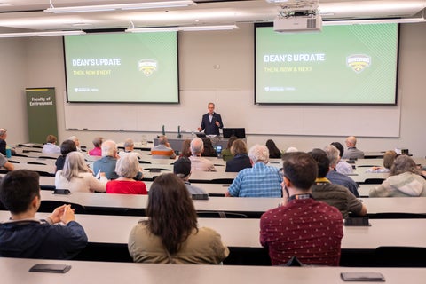 A male speaker in a dark suit stands at a podium addressing an audience seated in a lecture hall. Two large screens behind the speaker display "DEAN'S UPDATE THEN, NOW & NEXT" and the "HOMECOMING" logo. A "Faculty of Environment" sign is visible on the left wall.