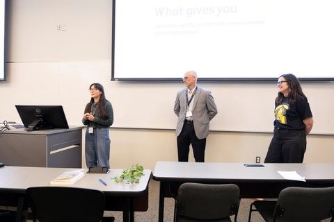 Three people stand in front of a projection screen in a classroom setting; one person stands at a podium to the left, presenting.