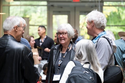 A group of people, including a woman in a black leather jacket wearing glasses and a lanyard, are gathered indoors.