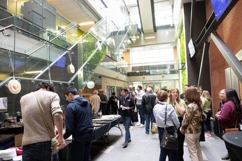 A group of people mingle in a modern atrium with a glass staircase and a vertical plant wall, some gathered around a food table.