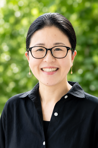 Smiling woman with black hair and glasses, wearing a black button-down shirt and gold drop earrings, outdoors.