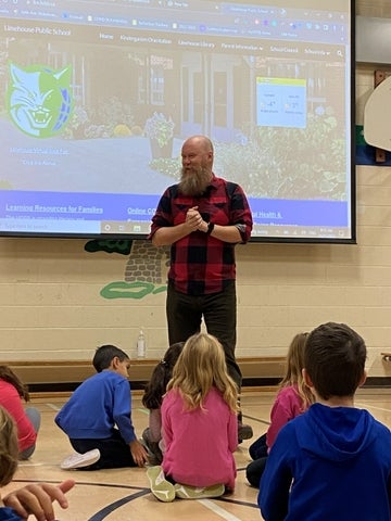 Bearded man in red plaid shirt speaks to seated elementary students in a school gym.