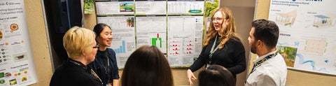 Individuals at a conference gathered din a circle discussing with academic posters shwoing bars, and graphs anad reserch findings in the bacground