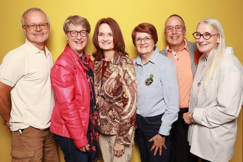 A group of six adults standing side by side against a yellow background, dressed in various styles and colors.