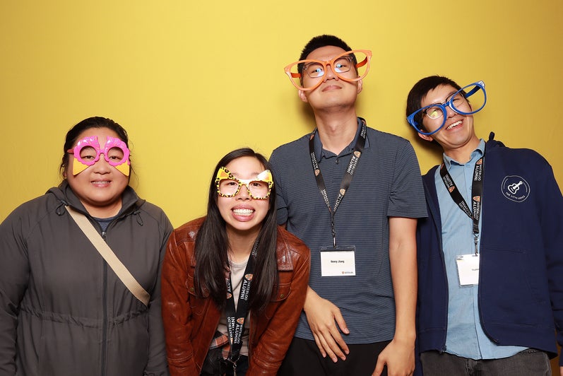 Four people with silly glasses smile in front of a yellow background.