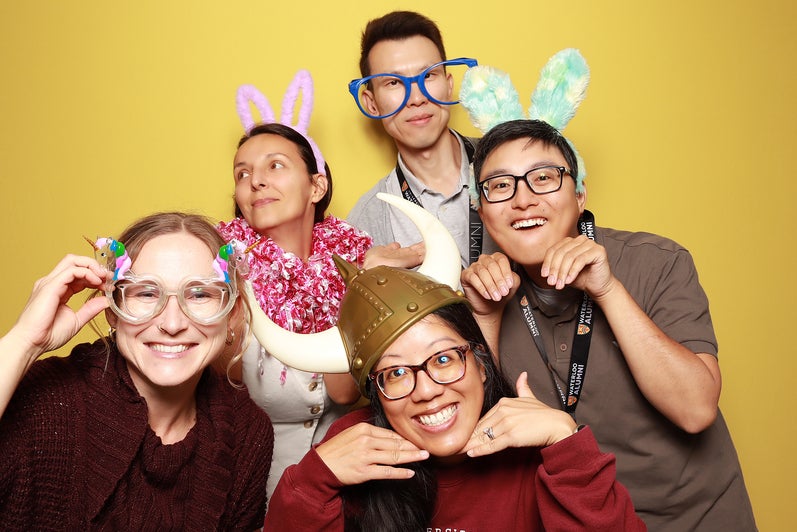 A group of five people pose with silly props in front of a yellow wall. 