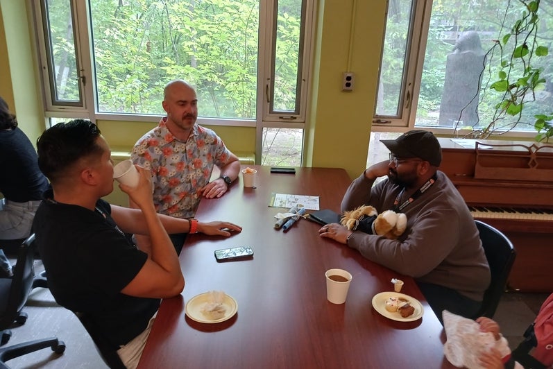 A group of three people sit at a table with coffees, talking. 