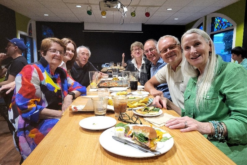 A group of people sitting at a long table in a restaurant, smiling and enjoying their meal.