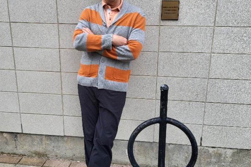 A man with glasses and a beard stands casually next to a circular signpost, wearing an orange and gray striped sweater, black pants, and sneakers, against a gray wall.