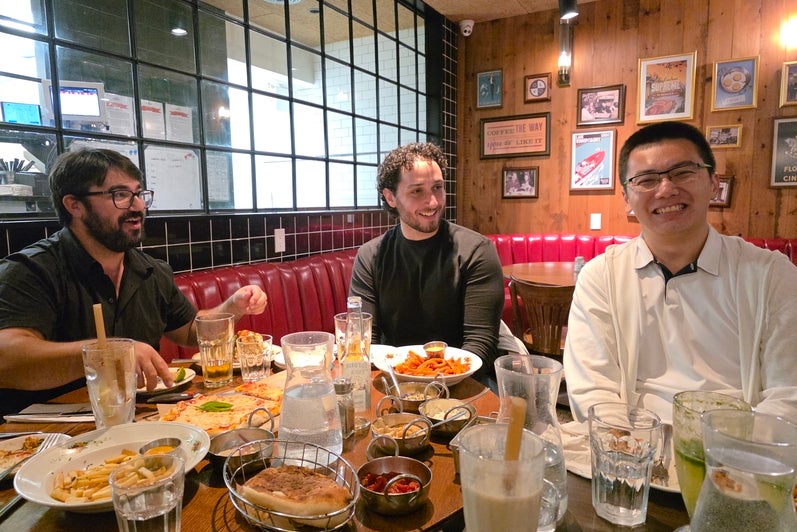 Three people are sitting at a restaurant table filled with food, smiling. 