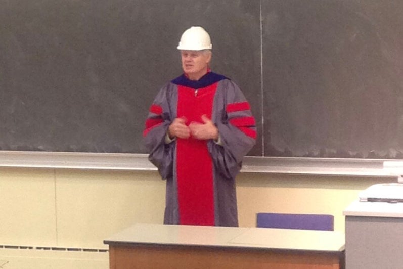 Man in a white hard hat and blue and red robes stands at the front of a classroom