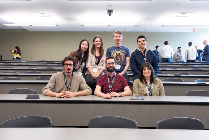 A group of seven people posing together in a lecture hall, with seats arranged in a grid behind them.