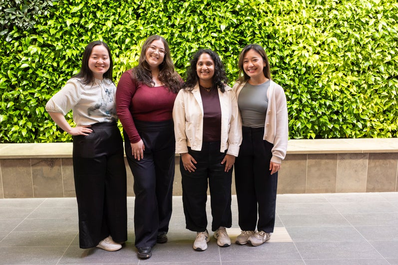 Four individuals stand in front of a green wall with leafy plants, smiling at the camera.