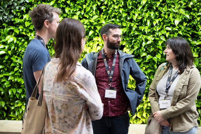 Group of four people stand together talking in front of green, plant wall. 