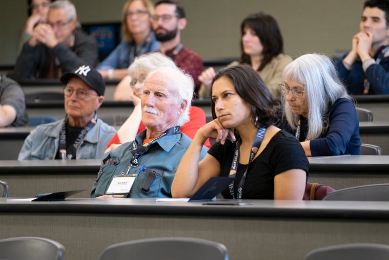 A diverse group of people sit in a lecture hall, listening. 