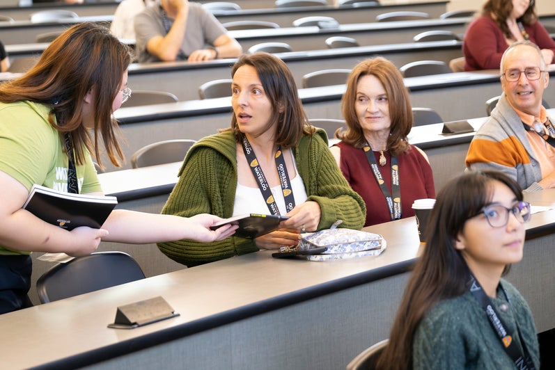 A diverse group of people sit in a lecture hall, with one being handed a book. 
