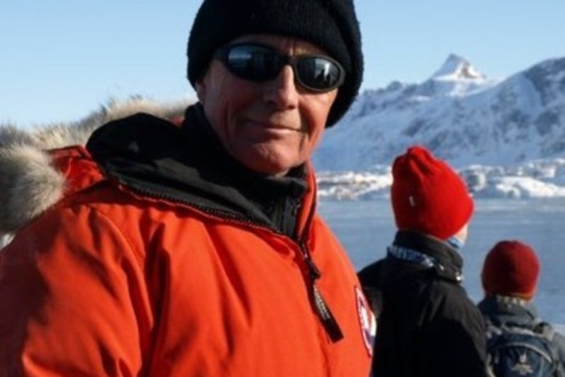 Man in red winter coat, black touque and sunglasses smiles with a wintery mountainous background.