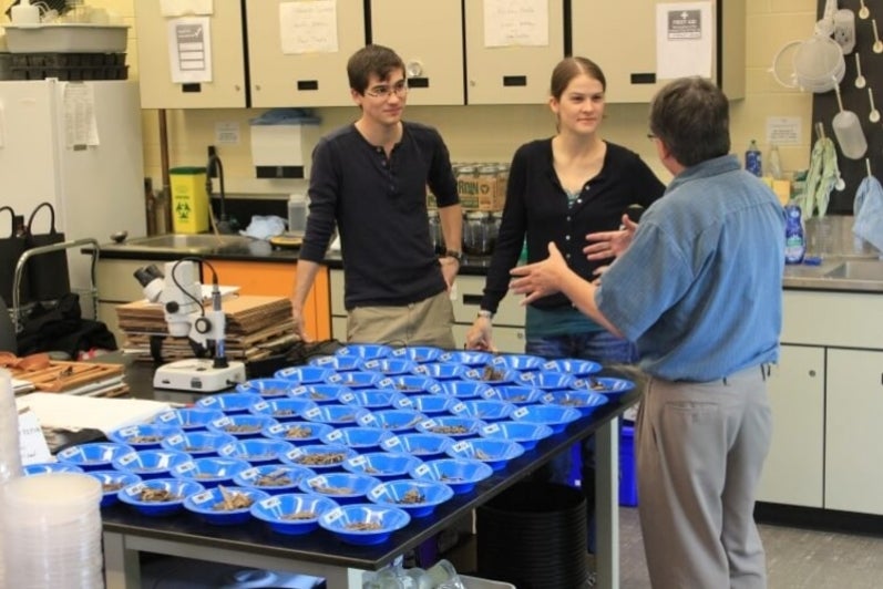 Professor in discussion with two grad students on far side of table, on which there is an array of blue trays full of samples