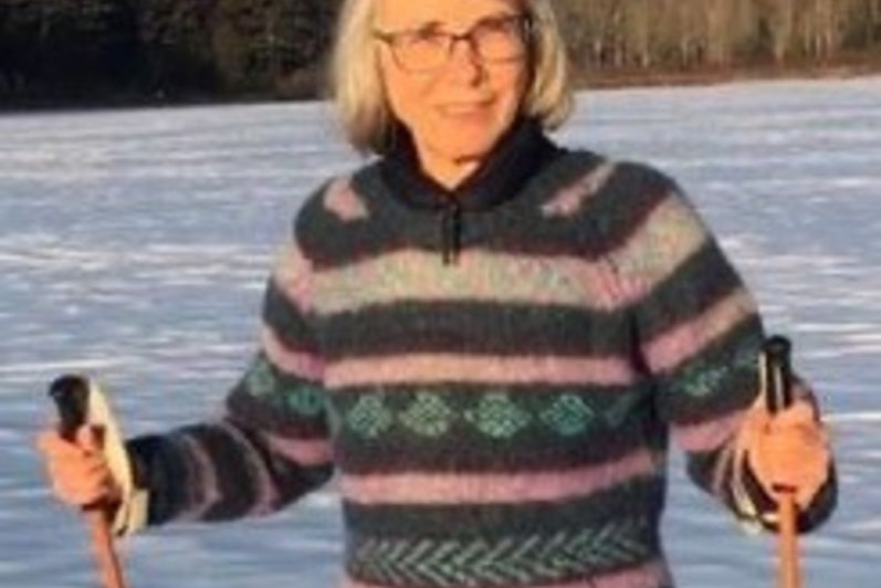 Woman with grey hair and glasses holds ski poles with snow in background.