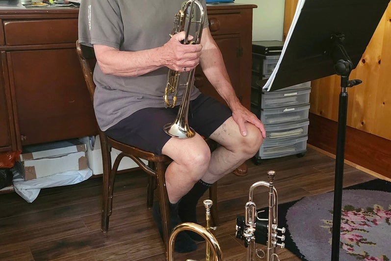 Man sits on stool with a trumpet in his hand and two trumpets on the ground. 