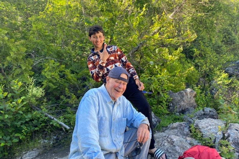 A smiling man in a light blue shirt and cap sits on rocks beside a younger person in a patterned shirt, surrounded by green trees.