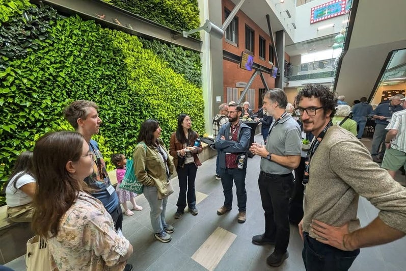 A group of seven people talking at a reception with high ceiling and a green plant wall. 