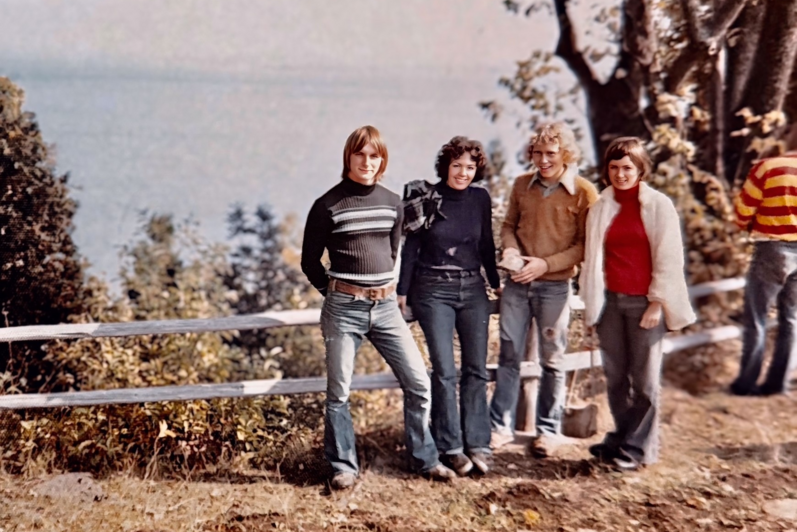 Four young people standing in a row outdoors by a wooden fence near a lake, dressed in 1970s-style sweaters, blue jeans, and a white fur jacket.