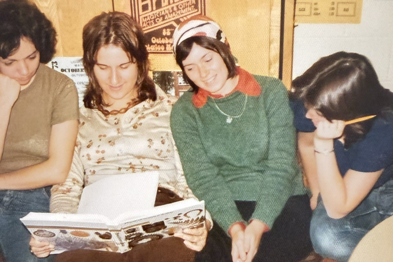 Four young women sit together, with two looking at an open book, one wearing a headscarf and green sweater, and another holding a yellow pencil.