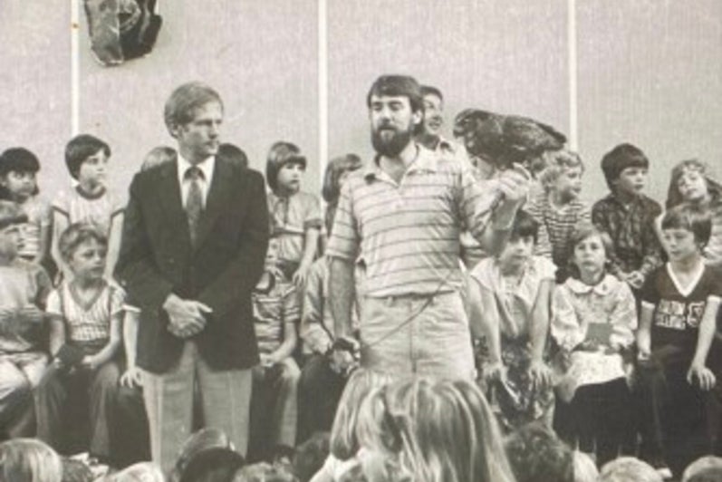 Black and white photo of man in striped shirt speaking in a group of children. 