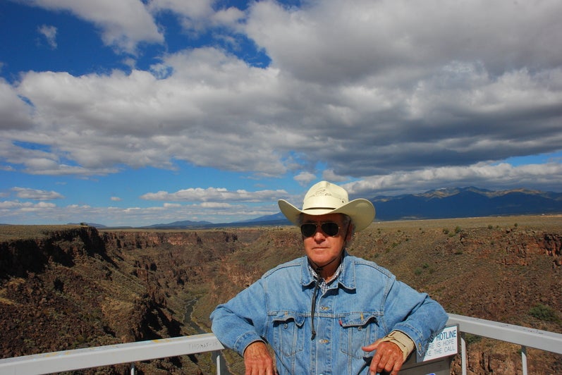 Man in denim shirt and white cowboy had leans against railing with prairies in the background. 