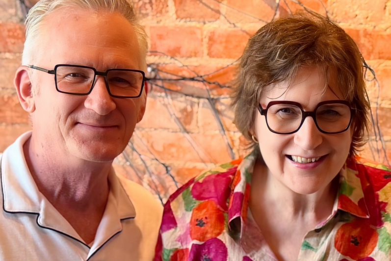 Man and woman, both in glasses smile in front of a brick wall.