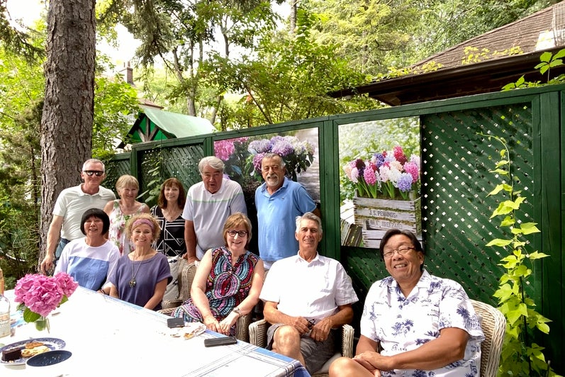 Group of people at a back yard table sit at table, with others standing behind them. 
