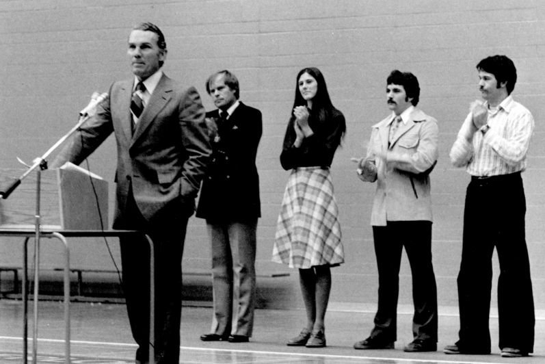 Man in suit speaks into microphone at podium while four students behind clap (black and white, 1977)