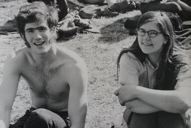 A black and white photo of a shirtless young man with sideburns and a young woman with long hair and glasses sitting together on the grassy ground.
